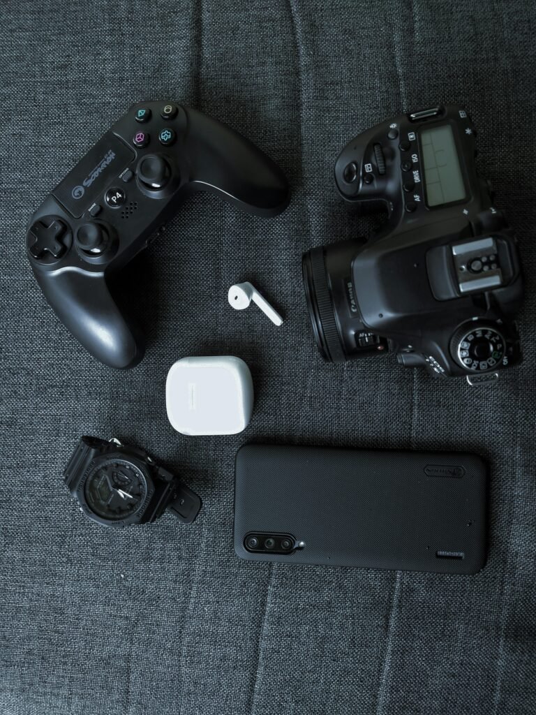 A collection of tech gadgets including a game controller, camera, smartphone, watch, and earphones on a fabric surface.